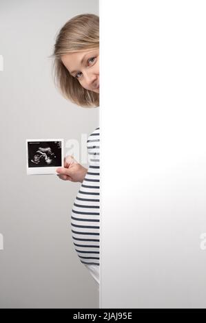 Femme enceinte avec une échographie se piquant hors du mur blanc. Bonne femme enceinte regardant l'appareil photo et montrant des ultrasons de sa photo de bébé. Banque D'Images