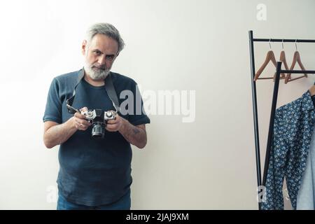 Le photographe mâle freelance caucasien âgé à cheveux gris tient son appareil photo et se tient dans son studio à côté du cintre à vêtements. Photo de haute qualité Banque D'Images