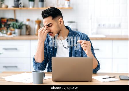Se sentir épuisé et fatigué pendant de longues heures de travail dans l'ordinateur portable. Frustré et triste, un indien ou un arabe a pris ses lunettes, fermé les yeux, movie son nez de pont, assis à son lieu de travail à son bureau à domicile Banque D'Images