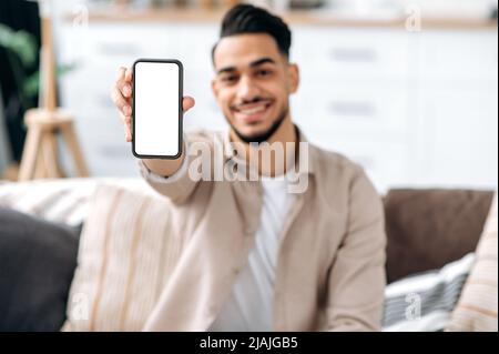 Un homme arabe ou indien souriant et défoqué, tenant un téléphone portable avec un écran maquette blanc vide en main, un espace pour la publicité ou la présentation, assis sur un canapé dans un salon intérieur Banque D'Images