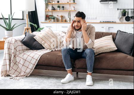 Triste homme arabe ou indien, asseyez-vous sur un canapé dans la salle de séjour, préoccupé par des problèmes personnels, triste par les mauvaises nouvelles, éprouvant la dépression, le désespoir, les maux de tête, la migraine, a besoin de repos et de soutien psychologique Banque D'Images