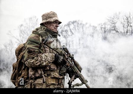 Chasseur de commando d'élite en uniforme de camouflage, armé d'un fusil de sniper, debout dans des nuages de fumée. Banque D'Images