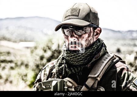 Soldat au visage sale fume une cigarette, portant des lunettes balistiques et une casquette de baseball. Banque D'Images