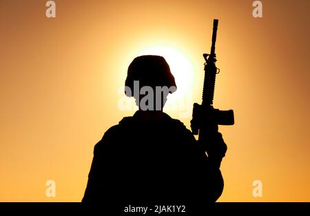 Silhouette d'un soldat debout avec un fusil d'assaut à la main contre un ciel de coucher de soleil. Banque D'Images