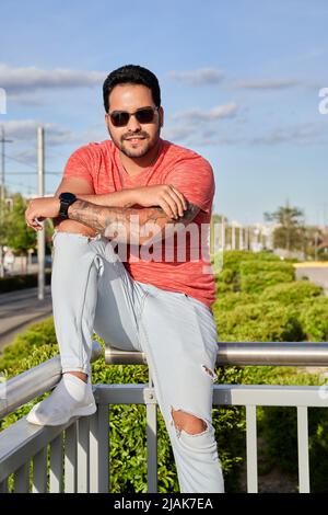 Latin homme avec des lunettes de soleil et des tatouages sur son bras regardant l'appareil photo et souriant tout en étant assis sur une main courante . Concept de style de vie urbain. Banque D'Images
