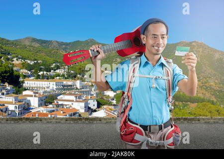 Homme asiatique avec un bonnet beanie et un sac à dos avec une carte et portant une guitare debout dans la rue avec un fond de cityscapes Banque D'Images