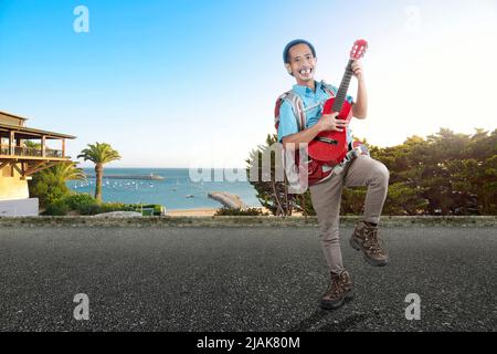 Homme asiatique avec un bonnet beanie et un sac à dos portant une guitare debout dans la rue avec une vue sur la plage Banque D'Images