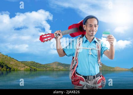 Homme asiatique avec un bonnet beanie et un sac à dos avec une carte et portant une guitare debout avec un fond de vue sur le lac et la colline Banque D'Images