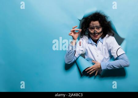 Spécialiste en biochimie insane avec cheveux désordonnés et visage sale grinçant la creepy tout en ayant le tube à essai en verre rempli de mélange chimique. Un chimiste fou avec un aspect délirant sur fond bleu. Prise de vue en studio Banque D'Images