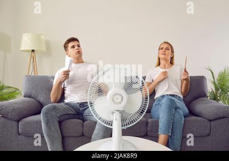 Jeune couple souffrant du temps chaud d'été et assis sur un canapé avec ventilateur électrique Banque D'Images