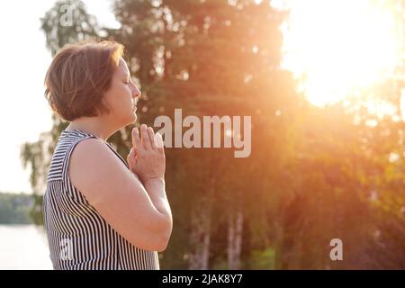 Une femme paisible tient ses paumes devant elle dans la prière contre les arbres de toile de fond sur le lac dans les rayons du soleil couchant. Méditer ou prier. La femme caucasienne croit en un pouvoir plus élevé Banque D'Images