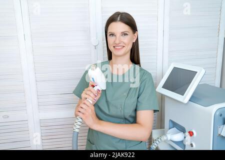 Gaie femme ajuste la machine d'enlèvement de cheveux de laser. Elle tient l'épilateur de la partie active dans ses mains et pose pour une photo. Il est situé dans un salon de beauté moderne. Soins du corps. Épilation laser des aisselles. Banque D'Images