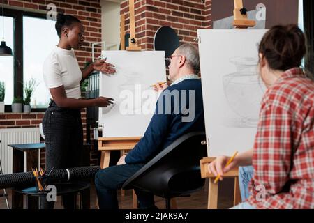 Jeune peintre enseignant expliquant la technique d'esquisse à un étudiant âgé pendant la leçon d'art. Groupe d'étudiants multiethniques dessin vase modèle sur toile blanche développement de compétences artistiques Banque D'Images