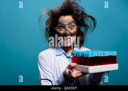 Chimiste fou et furieux avec plateau de verrerie de laboratoire avec tubes à essai remplis de substance liquide rouge. Scientifique en colère lunatique ayant des tubes de laboratoire en verre remplis d'échantillon sur fond bleu. Banque D'Images