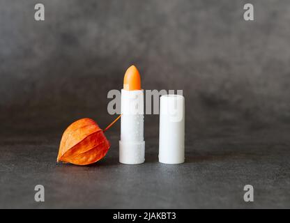 Tube blanc ouvert de rouge à lèvres hygiénique avec gouttes d'eau et physalis sec sur fond gris avec foyer sélectif. Copier l'espace pour le texte. Banque D'Images