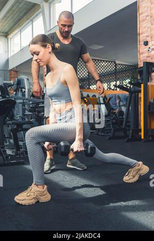 Prise de vue verticale en intérieur dans une salle de sport d'une jeune femme caucasienne à cheveux foncés dans une tenue de sport grise faisant une fente. Un homme européen chauve qui la soutient. Photo de haute qualité Banque D'Images