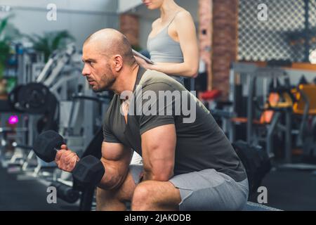 Photo en intérieur d'un bodybuilder masculin sans poils, qui reste concentré pendant l'haltérophilie, assis sur un banc dans un joli espace de fitness bien équipé. Photo de haute qualité Banque D'Images