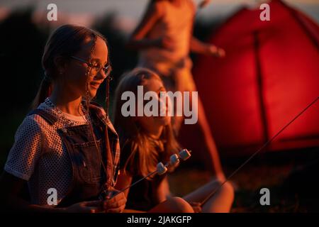 Deux adolescentes mangeant un repas rôti, s'amusant, assis près du feu de joie au crépuscule. Vue latérale de jolies filles qui s'accrochent des guimauves sur une brochette pour faire frire sur un feu de camp dans le camping. Concept de camping. Banque D'Images