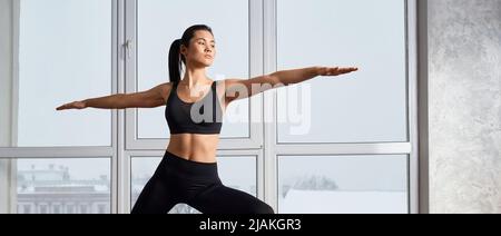 Vue latérale de la femme à la coupe flexible pratiquant le yoga dans la posture du guerrier II dans le spacieux studio de cours de yoga. Yogi fille avec queue de cheval restant dans Virabhadrasana II devant une large fenêtre. Concept de yoga asanas. Banque D'Images