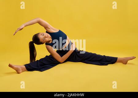 Jolie femme pratiquant l'asana de yoga avec des jambes qui s'étirent et se penchent d'un côté à l'intérieur. Une fille pieds nus avec des yeux fermés essayant d'atteindre la main au pied tout en faisant du yoga, isolée. Concept de l'exercice. Banque D'Images