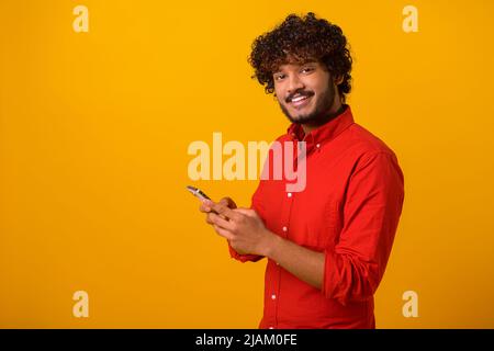 Portrait de l'homme barbu attrayant utilisant un téléphone portable avec une expression heureuse, accro au smartphone, SMS dans le réseau social. Studio d'intérieur isolé sur fond orange Banque D'Images
