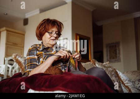 Active sénior rouge-cheveux retraité femme en lunettes a parlé pull-over tricoting, s'asseoir sur le canapé à la maison. Petite entreprise à la maison Banque D'Images