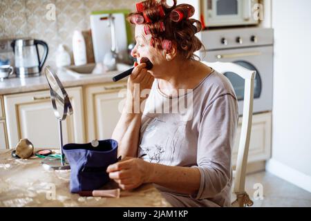 La vieille femme active âgée avec des rouleaux dans les cheveux prend soin d'elle-même le jour de son congé. Maquillage au pinceau, en regardant le miroir Banque D'Images