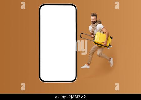 Application mobile pour les voyageurs. Un jeune homme à barbe enthousiaste bondissant avec une valise à bagages jaune près d'un immense smartphone avec écran vide, espace de copie, maquette. Offre vacances, vente Banque D'Images