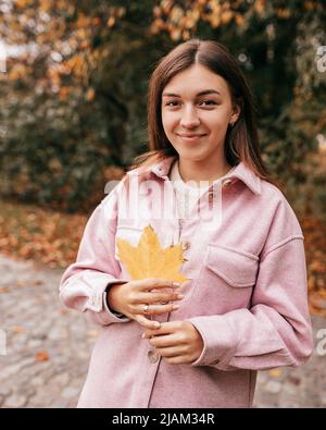 Jeune femme souriante aux cheveux longs bruns, en chemise rose chaude tenant la feuille d'érable d'automne, appréciant la marche dans le parc de la ville d'automne. Heure d'automne. Week-end Banque D'Images