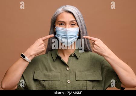 Sécurité pendant une pandémie, une épidémie, une grippe saisonnière. Femme âgée positive dans un masque médical sur son visage et pointe ses doigts vers le masque. L'appel à vous protéger Banque D'Images