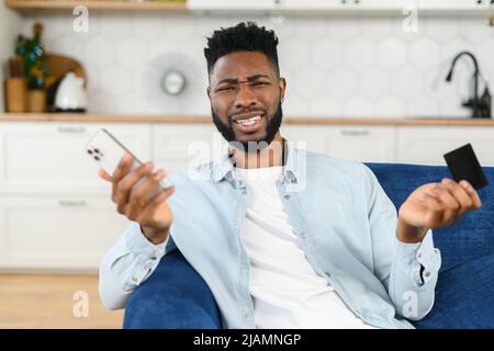 Un homme multiracial perplexe dans une chemise élégante et décontractée tenant un smartphone et une carte de crédit tout en exprimant un malentendu, répand ses mains dans l'incompréhension Banque D'Images