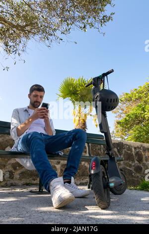 Le jeune homme regarde son smartphone assis sur un banc de parc à côté de son scooter électrique Banque D'Images