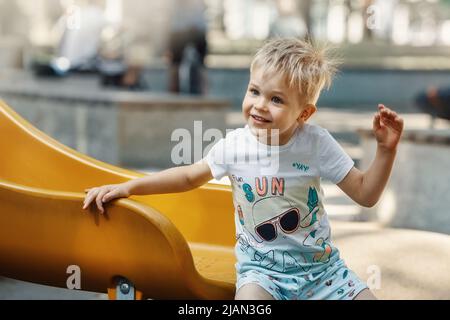 Un petit garçon joyeux, souriant et agréable sur l'aire de jeux sur un curseur jaune. Les cheveux de l'enfant sont électrifiés et picots. Banque D'Images