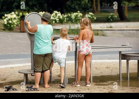 Petit garçon portant un short et un t-shirt blanc jouant avec l'eau du robinet public, ses amis grand garçon et fille pompant. Banque D'Images