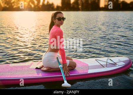 Belle jeune femme en lunettes de soleil et vêtements de sport flottant sur le paddle-board pendant le coucher de soleil incroyable sur le lac. Concept de modes de vie actifs et sains. Banque D'Images