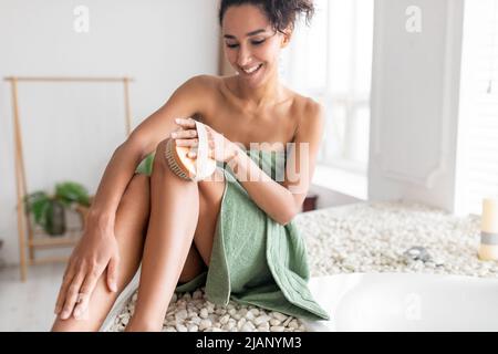 Belle jeune femme dans une serviette sèche se brossant les jambes, faisant anti cellulite ou massage lymphatique près du bain de bulles à la maison Banque D'Images