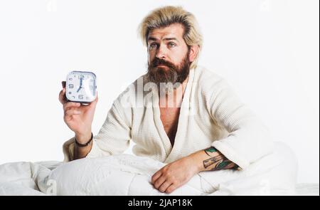 Homme à barbe endormi avec réveil dans le lit. Heure du matin. Eraillement. Banque D'Images