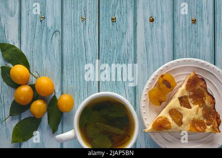 Un morceau de tarte aux prunes de cerise avec une tasse de thé à la menthe et des baies de prunes de cerise sur une table en bois peinte en bleu. Matin dans le jardin Banque D'Images