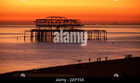 Silhouetted restes de la jetée ouest décrépit contre un ciel orange riche coucher de soleil. Brighton & Hove, Sussex, Angleterre, Royaume-Uni. Silhoueted formes de personnes sur la plage en premier plan. Banque D'Images