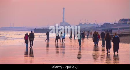 La foule de personnes marchant sur la plage au coucher du soleil. Ciel rose sombre. Marée basse, Brighton & Hove, East Sussex, Angleterre, Royaume-Uni. En regardant vers l'ouest depuis Hove vers Shoreham et la centrale électrique à Shoreham Harbour. Banque D'Images