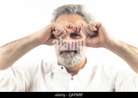 Studio tourné sur fond blanc d'un homme européen attrayant dans son 60s faisant un signe de coeur avec ses doigts sur son visage. Mise au point au premier plan. Photo de haute qualité Banque D'Images