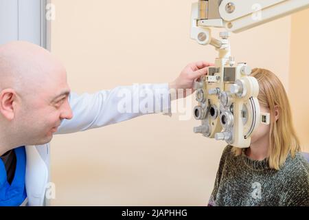 Un ophtalmologiste mâle vérifie la vue d'une jeune fille à l'aide d'un phoroptère. Traitement de la vision. Banque D'Images