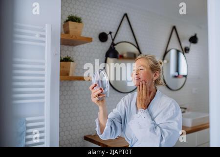 Belle femme senior en peignoir regardant le miroir et appliquant la crème naturelle du visage dans la salle de bains, concept de soins de la peau. Banque D'Images