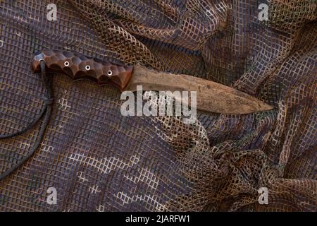 Un couteau militaire repose sur un filet de camouflage, armes bordées Banque D'Images