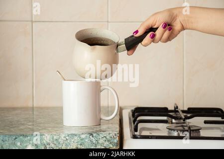 La main d'une femme verse le café d'un turk dans une tasse sur la table de la cuisine, préparant le café du matin dans la cuisine Banque D'Images