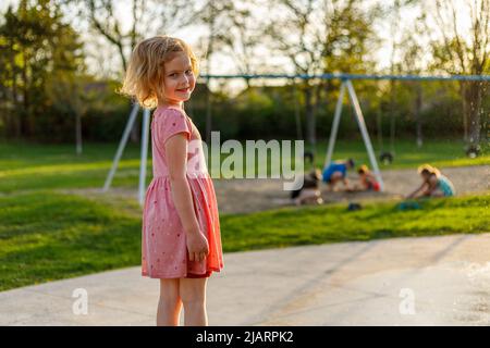 Une petite fille souriante et des enfants qui jouent dans le sable près des balançoires dans le parc lors d'une soirée ensoleillée d'été Banque D'Images