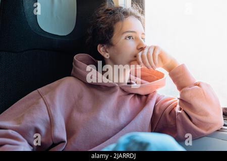 l'adolescent solitaire pensif est assis dans le train près de la fenêtre, se reposant la tête sur la main, regarde la fenêtre, se pliant les genoux. Long trajet en train, jambes gonflées, voyage en Europe Banque D'Images