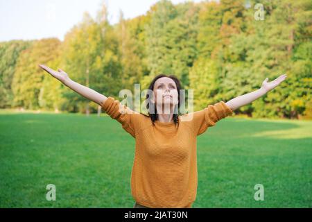 Jolie femme debout dans un parc naturel de printemps avec de larges bras ouverts relevés dans un chandail jaune clair. Belle herbe verte fraîche et feuillage par temps ensoleillé Banque D'Images