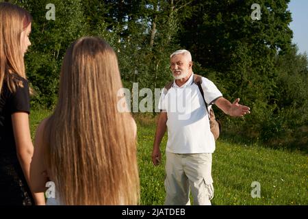 Vue latérale d'un homme avec des cheveux gris et une barbe qui parle avec des filles en bois. Vieil homme sérieux expaining, pointant à la main vers deux jeunes filles à l'extérieur, concept de repos actif. Banque D'Images