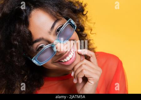 femme afro-américaine en lunettes de soleil bleues touchant le menton et regardant la caméra isolée sur le jaune Banque D'Images